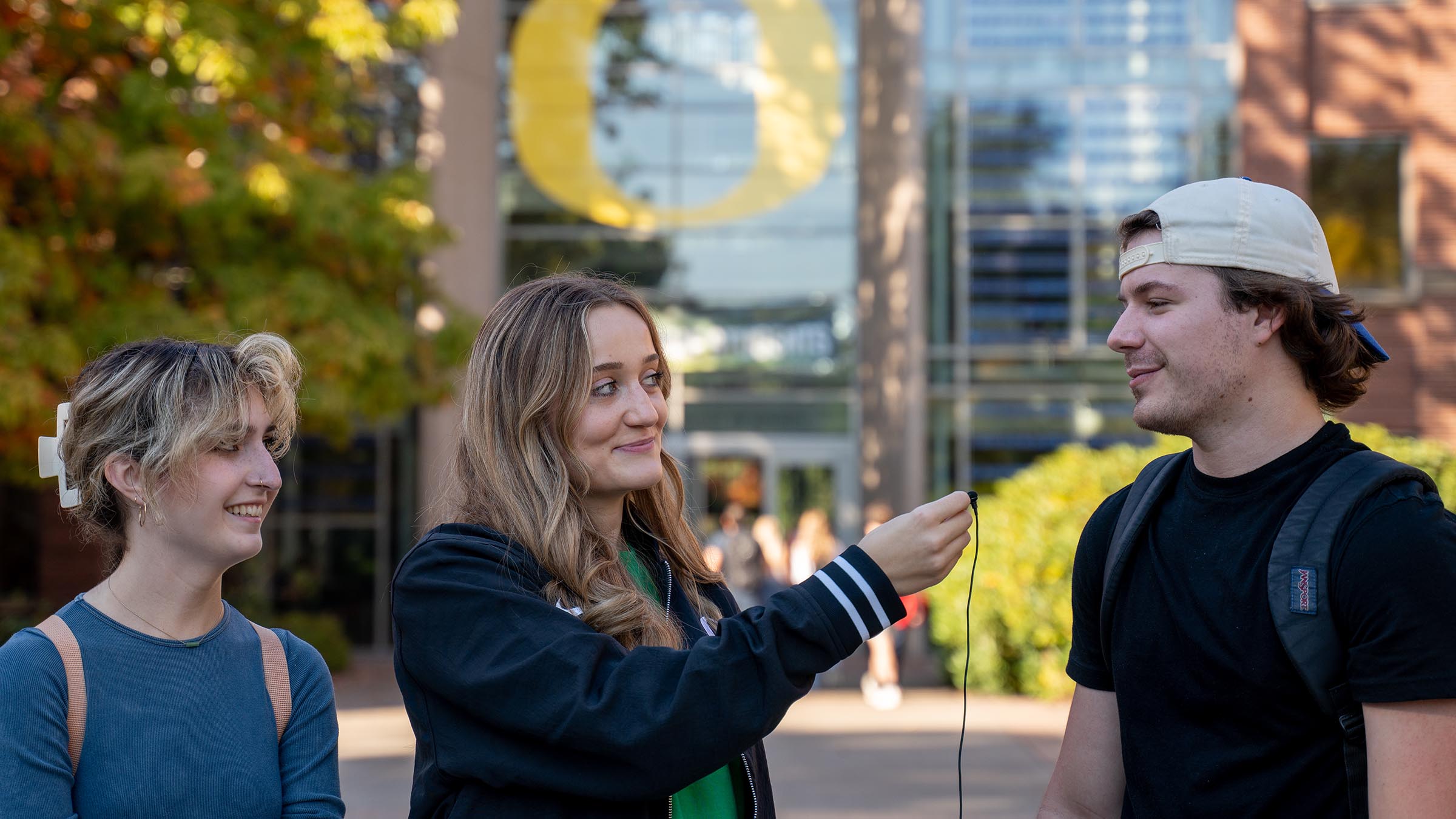 Student interviewing classmates on campus in front of the Lillis Business Complex